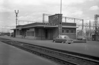 Image shows railway tracks and a station with a few automobiles parked beside it.