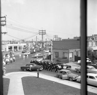 Image shows a street view at the intersection. There are many parked cars and people dressed fo…