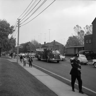 Image shows a street view. There are some vehicles participating and some people walking on the…