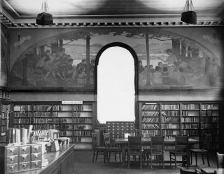 Image shows an interior of the library branch with book stacks along the walls, the staff desk …