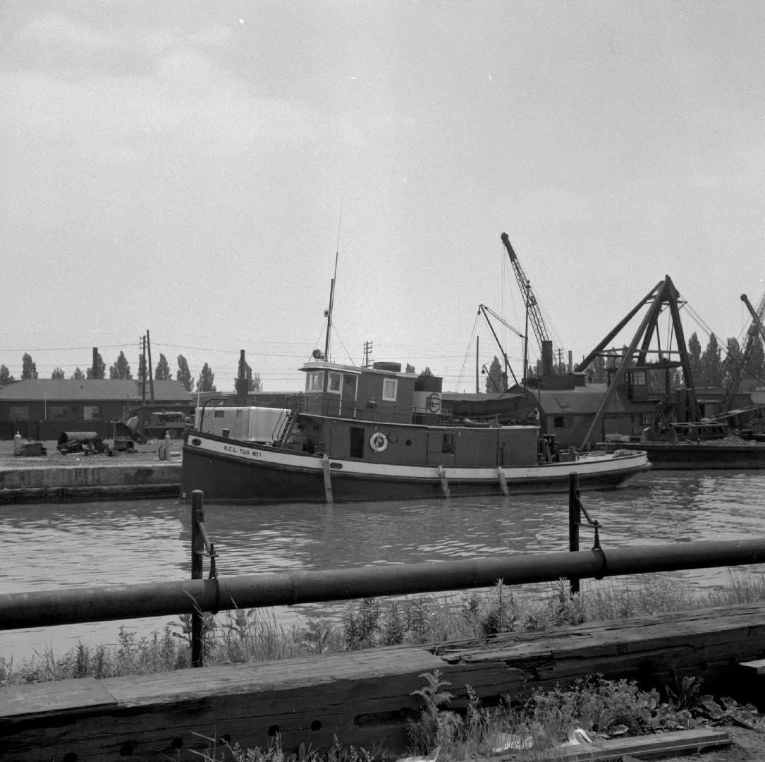 Image shows a docked tugboat with some cranes in the background.