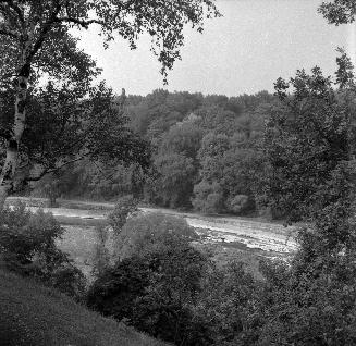 A photograph of a river, with a steep hill in the foreground and heavily wooded areas on both s…