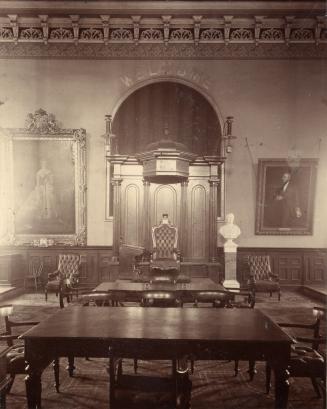 Image shows an interior of the hall. There are some desks and chairs, a few big pictures hangin…