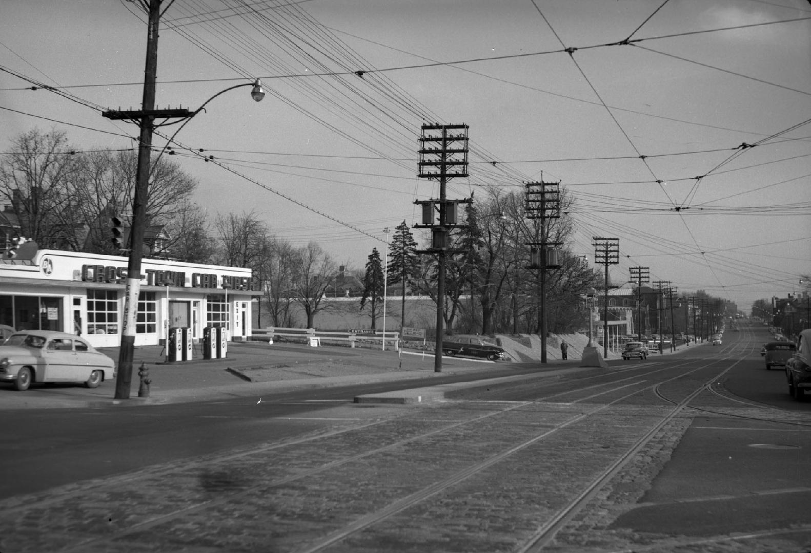 Image shows a street view with trees on both sides. There is a car parked on the left.