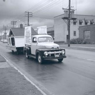 Image shows a car participating in the parade that is pulling a small house and has some poster…