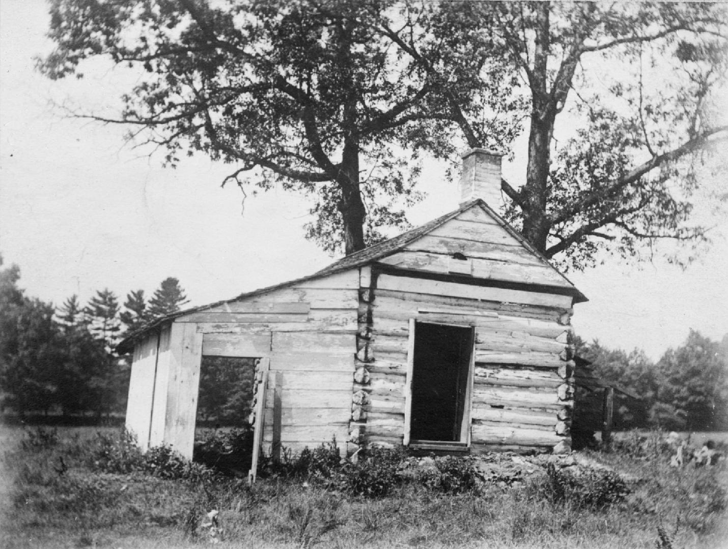 Image shows a log cabin. There are a few trees behind it.