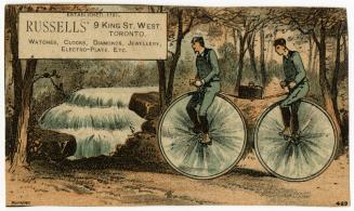 Two men riding on a tandem bicycle with very large wheels (like a penny farthing). They are rid…