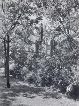 Image shows a very limited view of a house as there are a lot of trees and bushes in front of i…