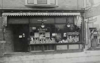 Image shows the front of a drug store that reads &quot;N. L. McMillan Druggist&quot;.