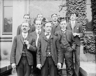 A formal group portrait of 8 men in three-piece suits and ties standing outdoors, on a small st…