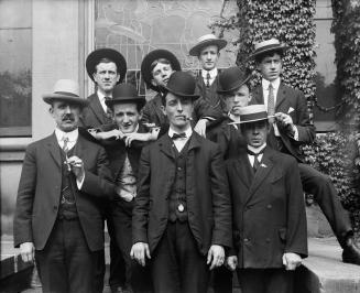 An informal group pose: 9 men in three-piece suits, and hats, posed on a short outdoor staircas…