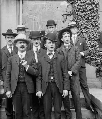 A formal, posed group portrait of 8 men, wearing hats and three-piece suits. They are posed on …