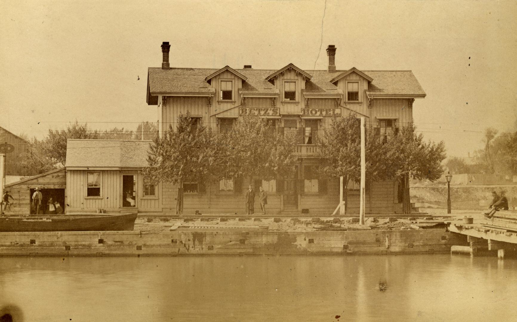 Image shows a waterfront three storey hotel with some trees in front of it.