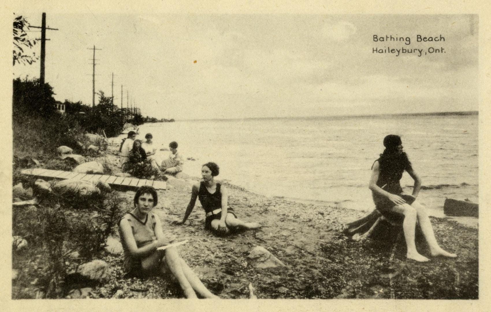 Photograph shows ladies in bathing suits sitting on a very rocky beach.