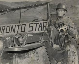 A man in army fatigues and a helmet sits, holding an old-fashioned camera, in an open jeep with…