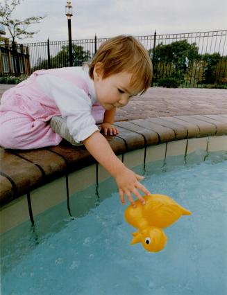 A toddler in a pink outfit leans precariously over the edge of a pool, reaching for a rubber du…