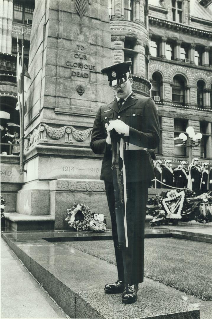 A man in uniform stands, with bowed head, in front of the cenotaph at Old City hall, which read…