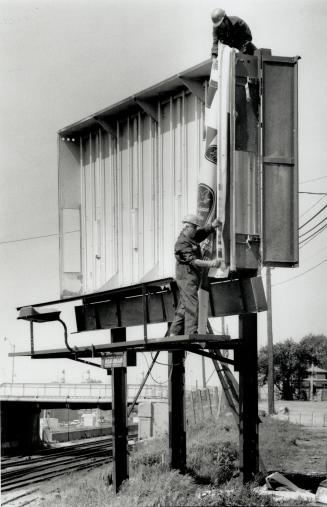 Easy as one, two three . . . Francis Andrade, bottom, and Louis Guinho, top, install a new display on a transparency-type billboard measuring 3 metres(...)