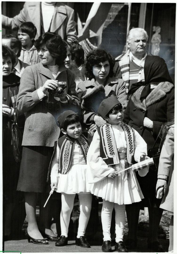 Two children dressed in white pleated skirts and tights, smocked vests and black caps, stand in…