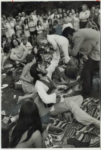 A bearded man with long hair sits on a blanket on the grass, holding a small baby in the air wi…