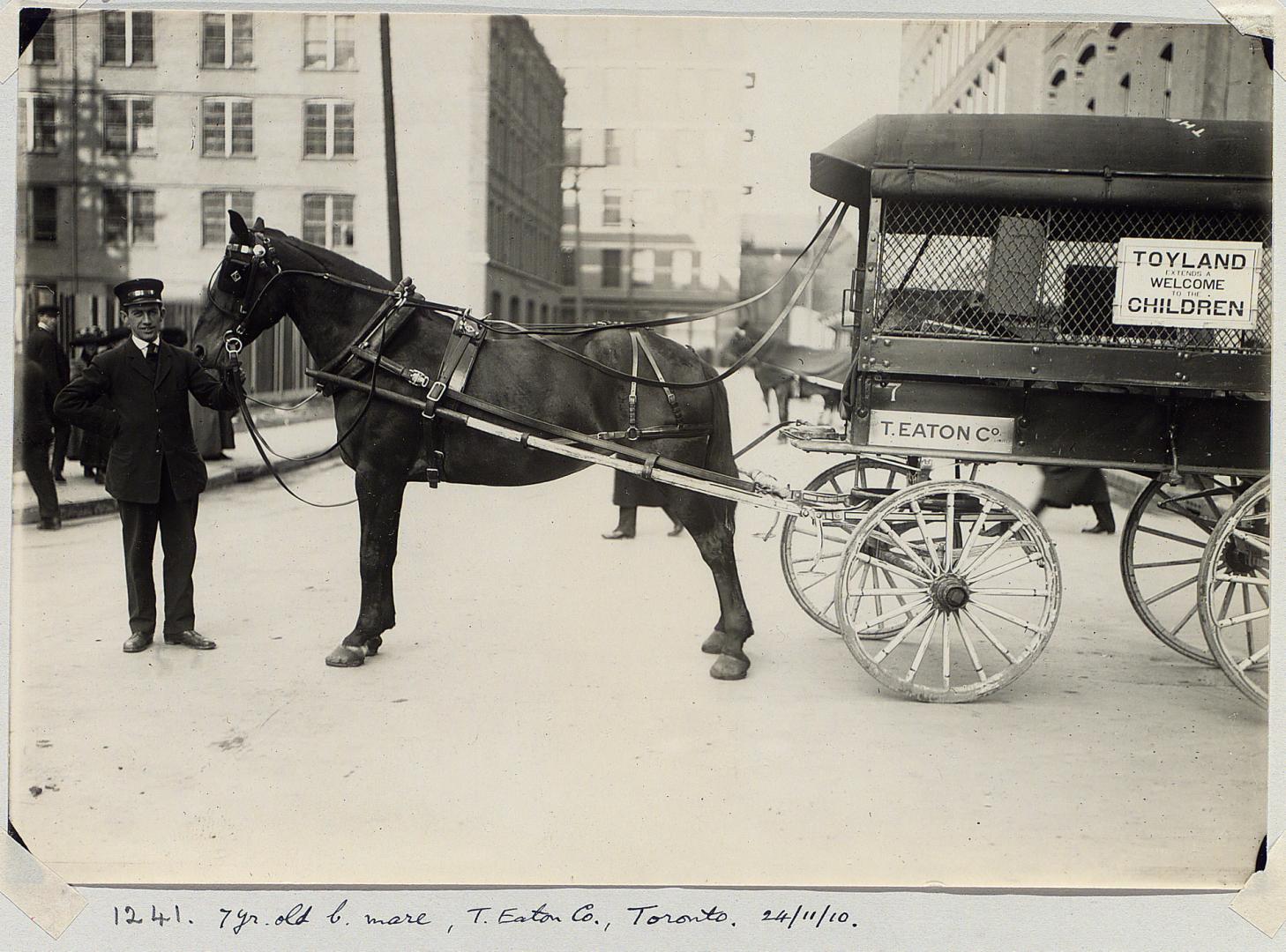 Eaton's delivery agent holding bridle of a horse harnessed to a wagon that says &quot;Toyland w…