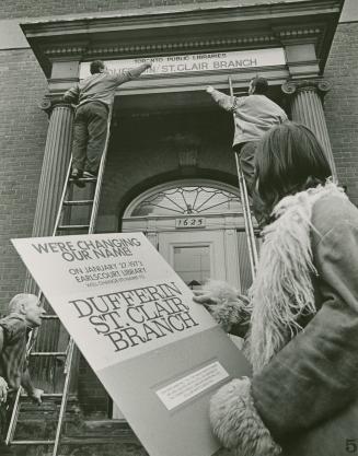 Woman watches two men on ladders install a library sign above entrance reading 'Toronto Public …