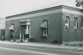 Street view of one-story brick building. Awnings cover arched windows on either side of the ent…