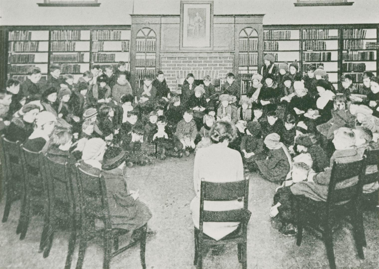 A group of children are gathered around a woman seated on a chair facing them. Shelves of books…