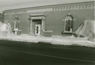 Façade of a one-story brick building with arched windows either side of a entrance flanked by p…