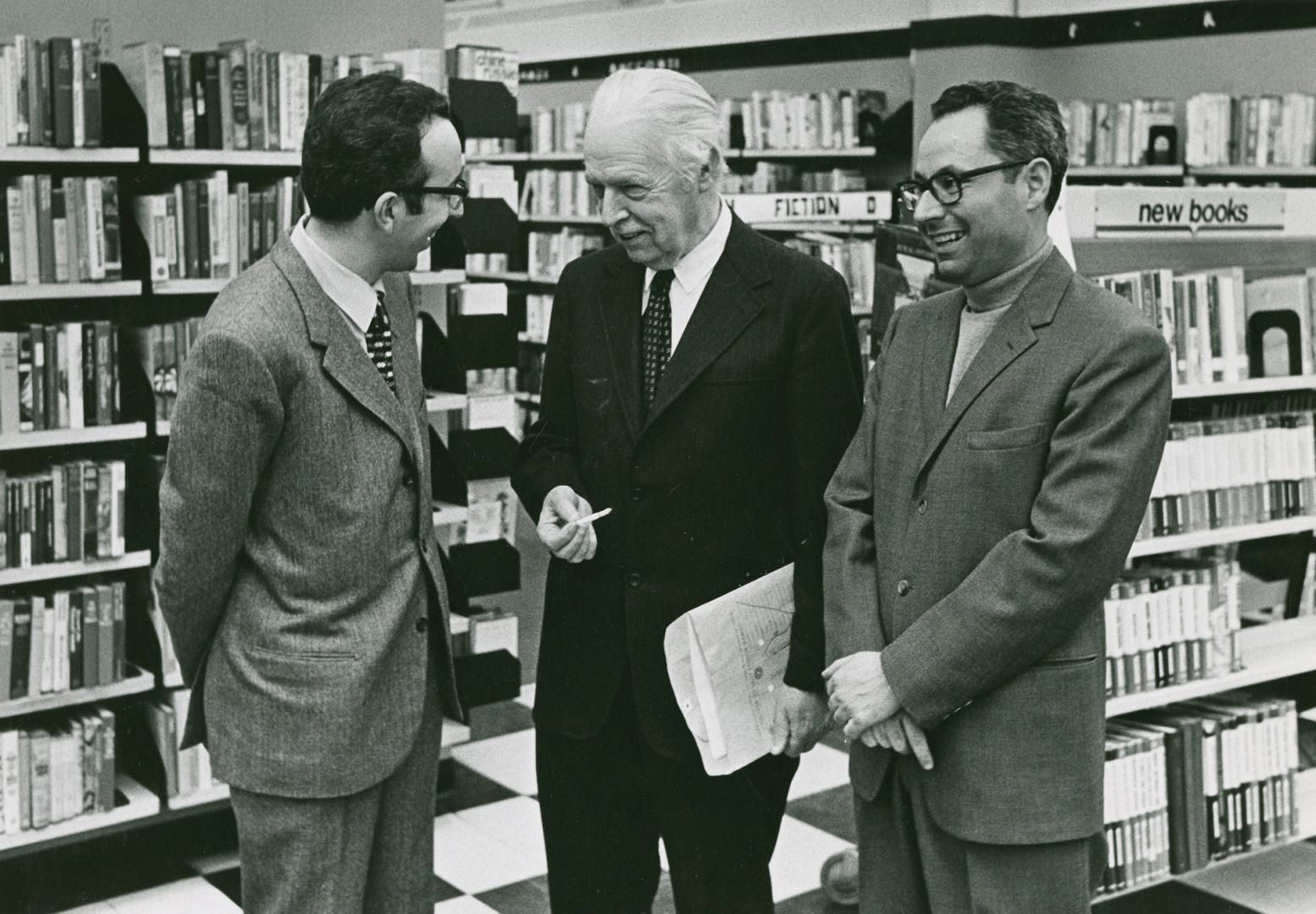 Three men in suits, one with papers in hand, congregate in front of a book shelf with a sign re…