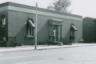 Street view of one-story brick building. Awnings cover the windows either side of an entrance f…