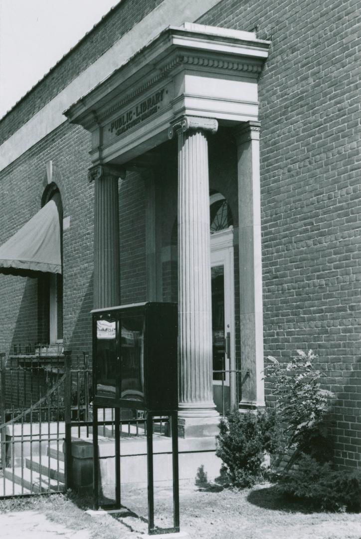 Side view of library entrance flanked by cement pillars . A black display case stands right of …