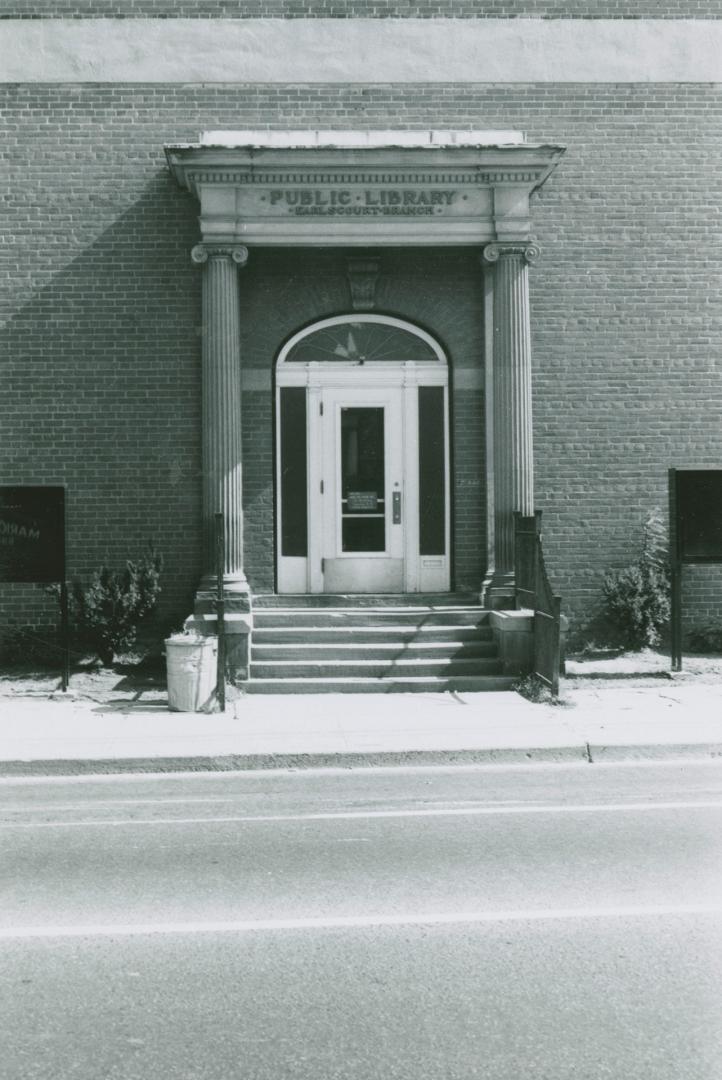 Front entrance of library. Steps lead to an arched door flanked by cement pillars. Sign above d…