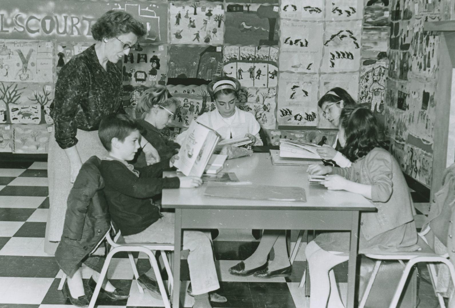 A woman stands near a table where children are reading. The surrounding walls are covered with …