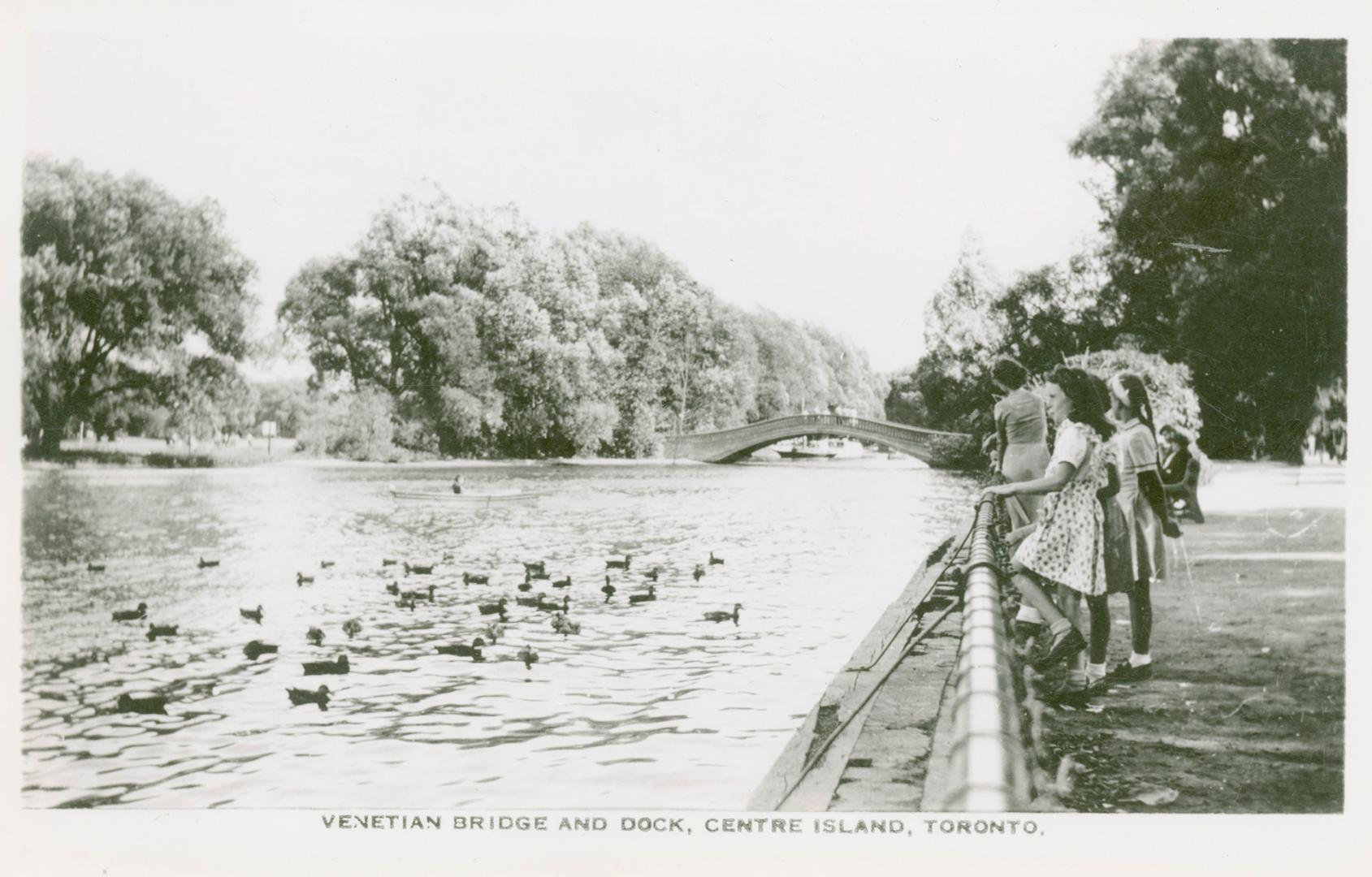 Black and white picture of a group of three girls standing on a dock watching ducks swimming in…