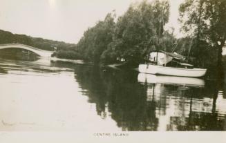 Black and white picture of a boat in a lagoon and bridge and trees in the background.