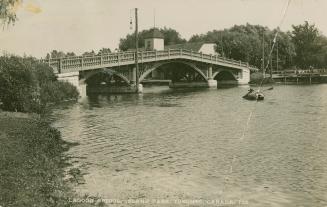 Photo of a bridge over a lagoon with a rowboat and trees in the background. 