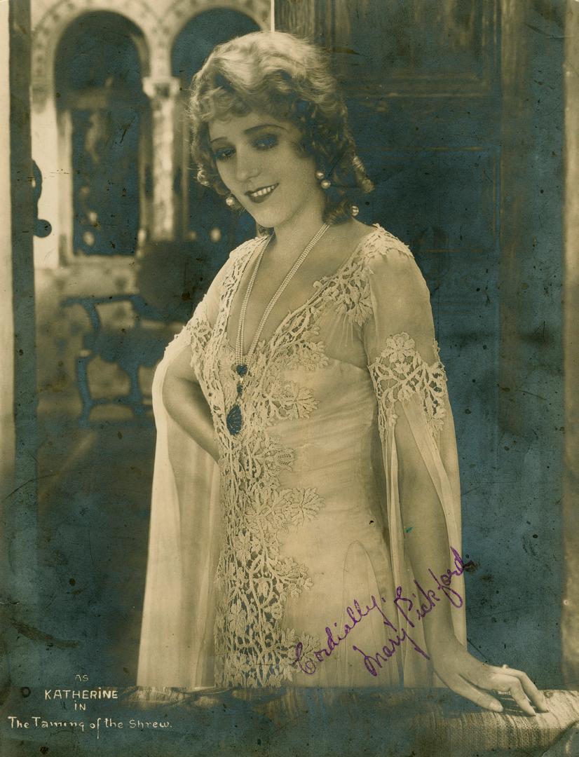 Mary Pickford in a white lace dress standing on the set of a stage.