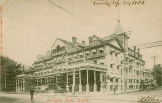 Black and white picture of a four story hotel with people and vehicles outside of it.