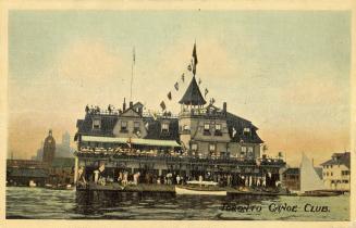 Colorized photograph of a large building beside a lake with many people standing on the dock in…