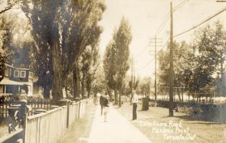 Black and white picture of a wooden boardwalk in front of cottages with three people looking at…