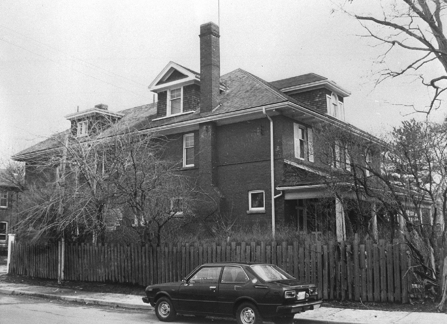 A photograph of a two-story house with a fence alongside it and a car parked in front of the fe…