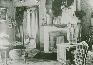 A photograph of the interior of a house, with a fireplace, chairs and other pieces of furniture…