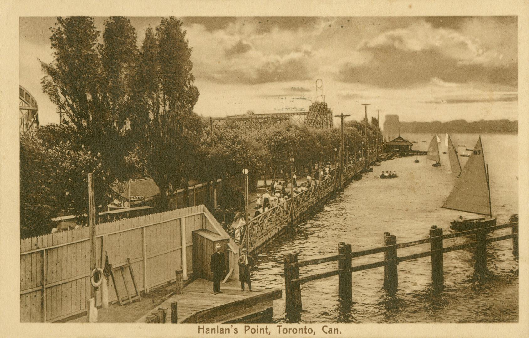 Black and white photo postcard of the boardwalk at Hanlan's Point, filled with people and showi…