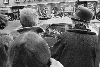 A photograph of a group of people standing on a sidewalk, with a car passing by.