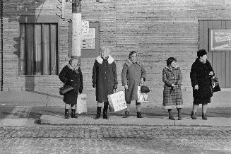 A photograph of five women standing on a sidewalk near a traffic light.