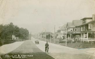 Black and white photograph of a police officer on a bicycle on a street with houses. Motor car  ...