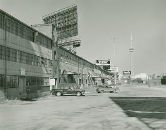 A photograph of a factory, with pickup trucks parked in a lot and the CN Tower visible in the b…