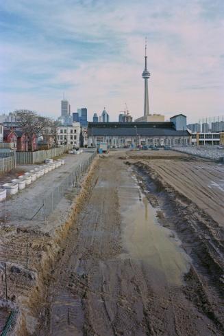 A photograph of a muddy field surrounded by houses on two sides, with skyscrapers and the CN To…