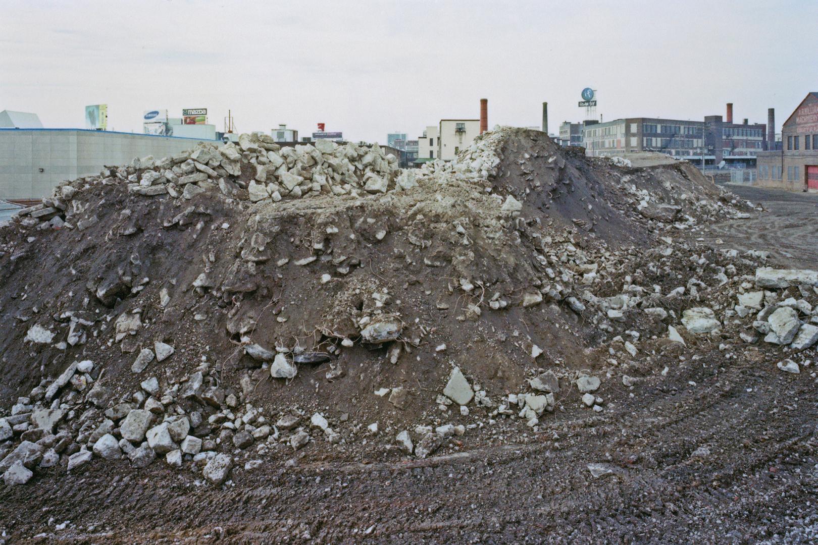 A photograph of a pile of dirt and construction rubble in the middle of a muddy field, with hou…